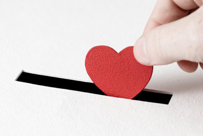 A hand placing a red textured heart into a black slit on a white surface, symbolizing inserting love or affection.