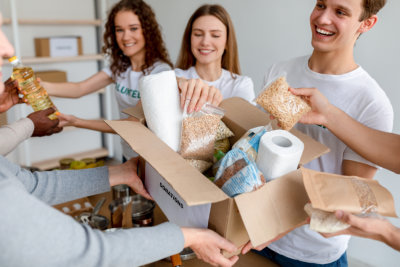 Group of cheerful young volunteers packing food and supplies into cardboard boxes in a bright room.